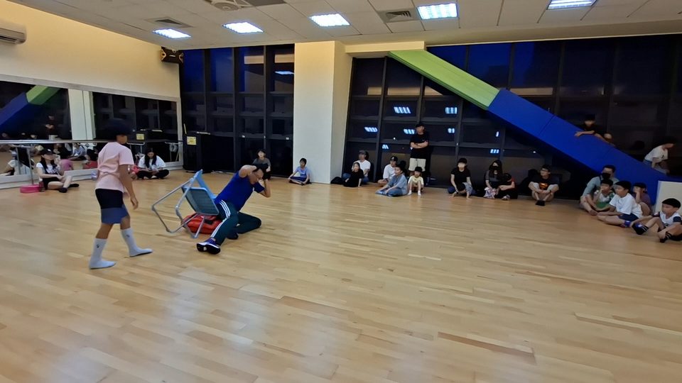 教練在活動室示範用折疊椅擋在身前,孩子們沿牆坐著看